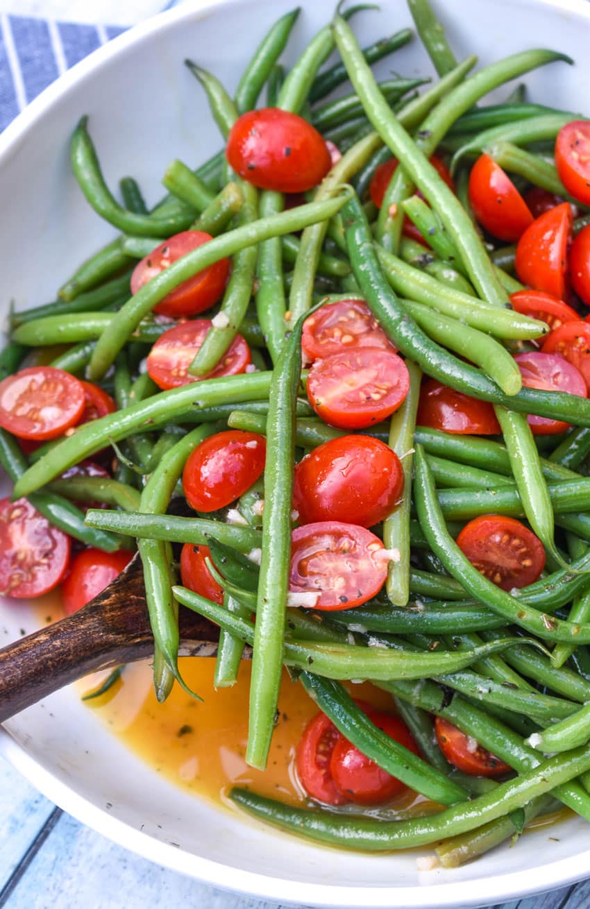 a wooden spatula scooping green bean and tomato salad out of a white serving bowl