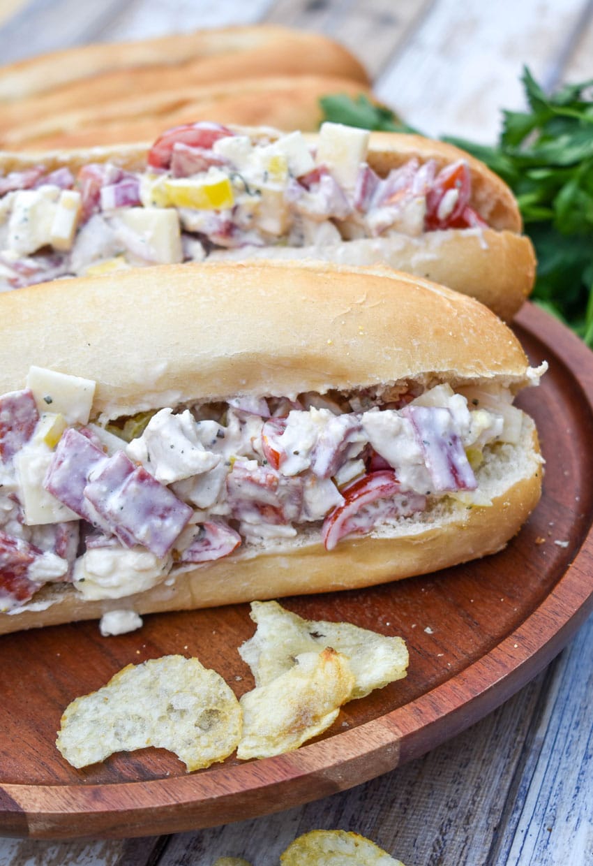 two italian grinder chicken salad sandwiches on a wooden plate with potato chips on the side