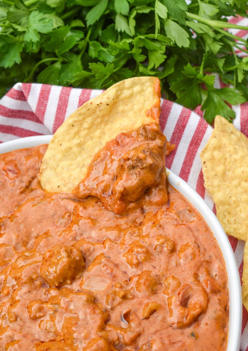 A TORTILLA CHIP TOPPED WITH THE BEST HOMEMADE CHILI CHEESE DIP RESTING ON THE EDGE OF A WHITE SERVING BOWL