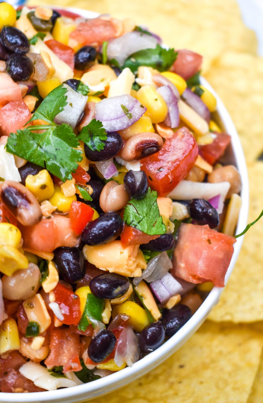 a close up of cowboy caviar dip in a white bowl surrounded by tortilla chips