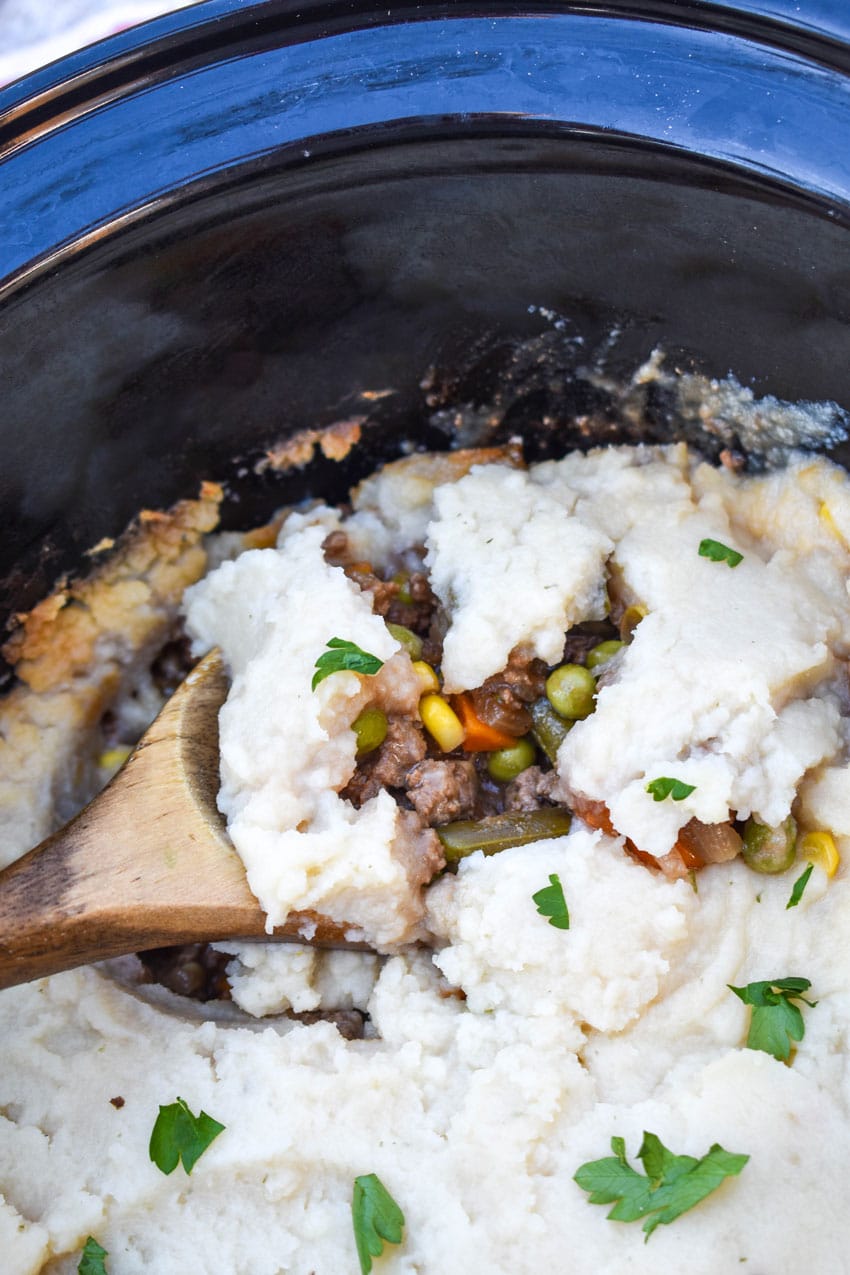 a wooden spoon scooping mashed potato topped shepherd's pie our of the black bowl of a slow cooker