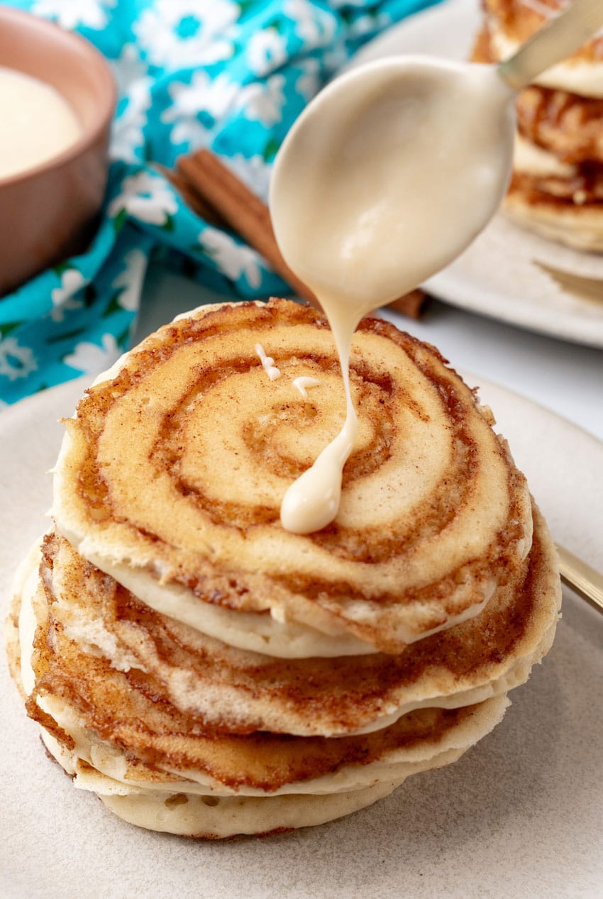 cream cheese icing being spooned over a stack of cinnamon roll pancakes