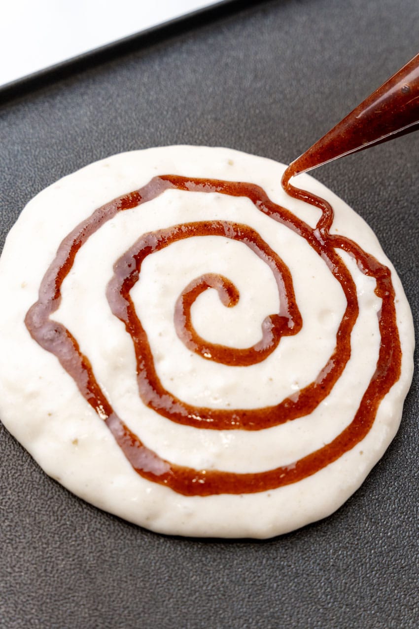 cinnamon sugar swirl being piped onto pancake batter on a hot griddle