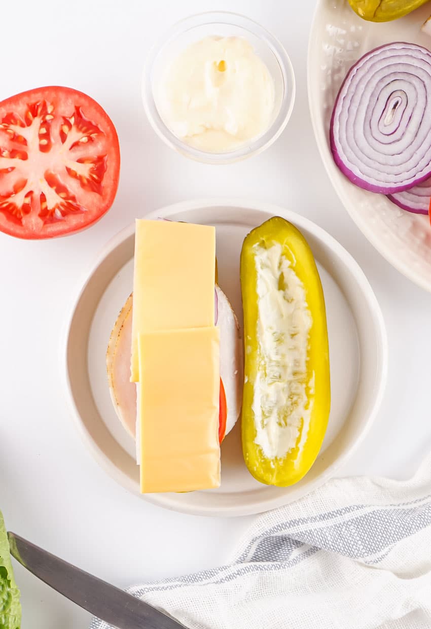 sliced turkey and layered veggies and sliced cheese on a mayo slathered dill pickle on a white plate