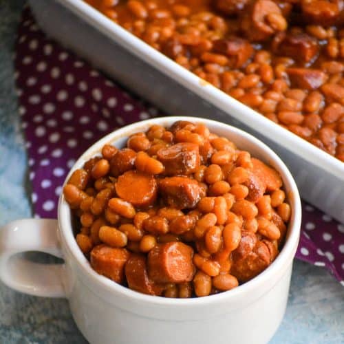 hotdogs and beans casserole in a white mug next to a white baking dish