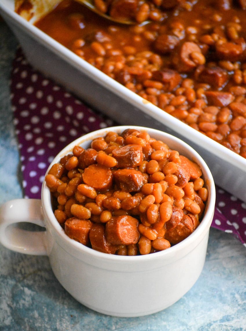 hotdogs and beans casserole in a white mug next to a white baking dish