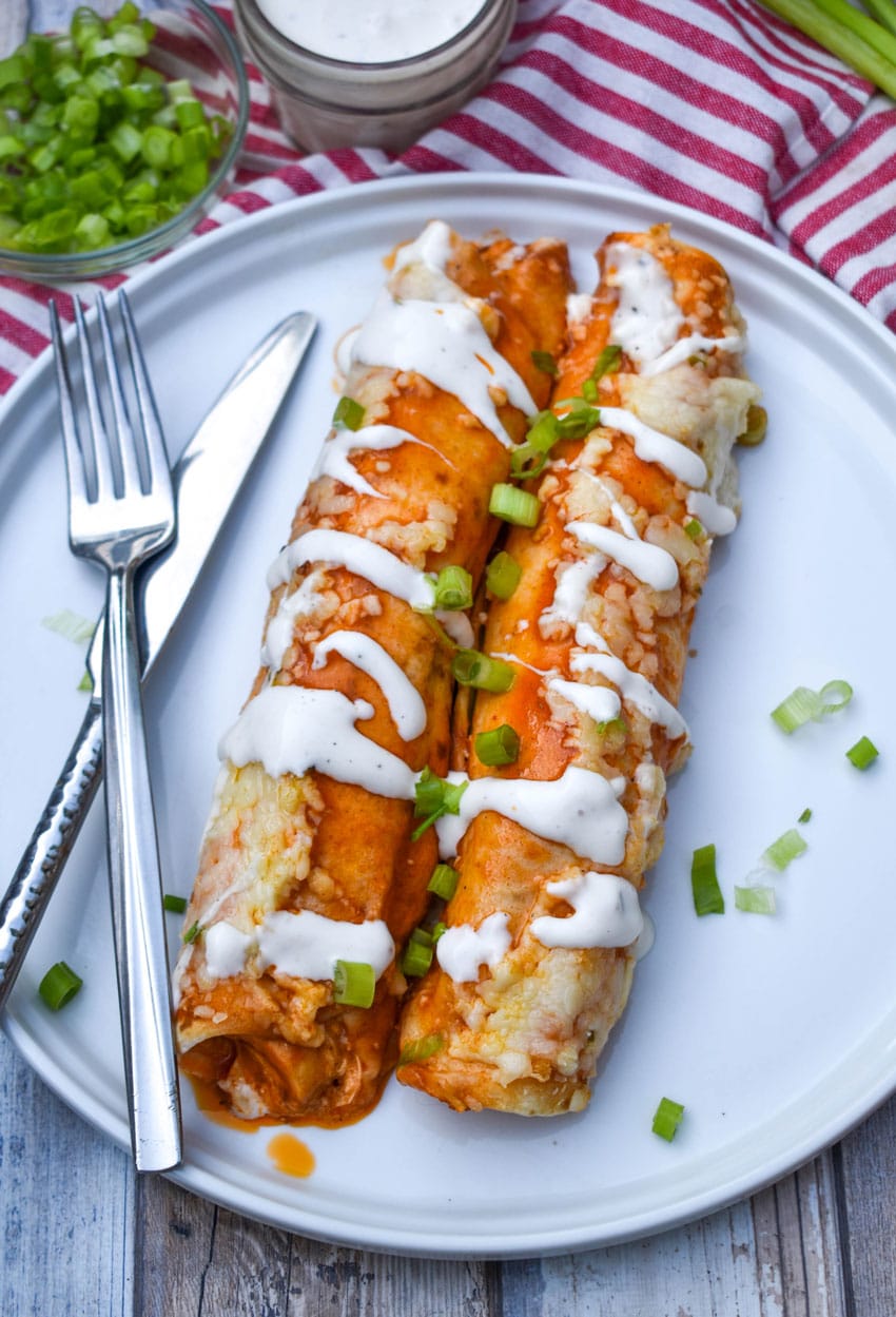 two buffalo chicken enchiladas topped with a drizzle of ranch dressing and sliced green onions on a white plate