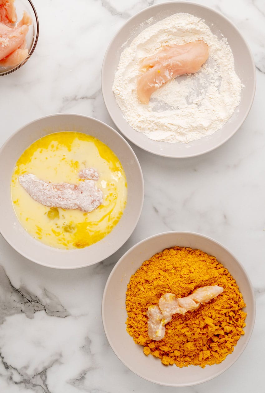 chicken strips being dipped in three shallow bowls in various stages of the breading process