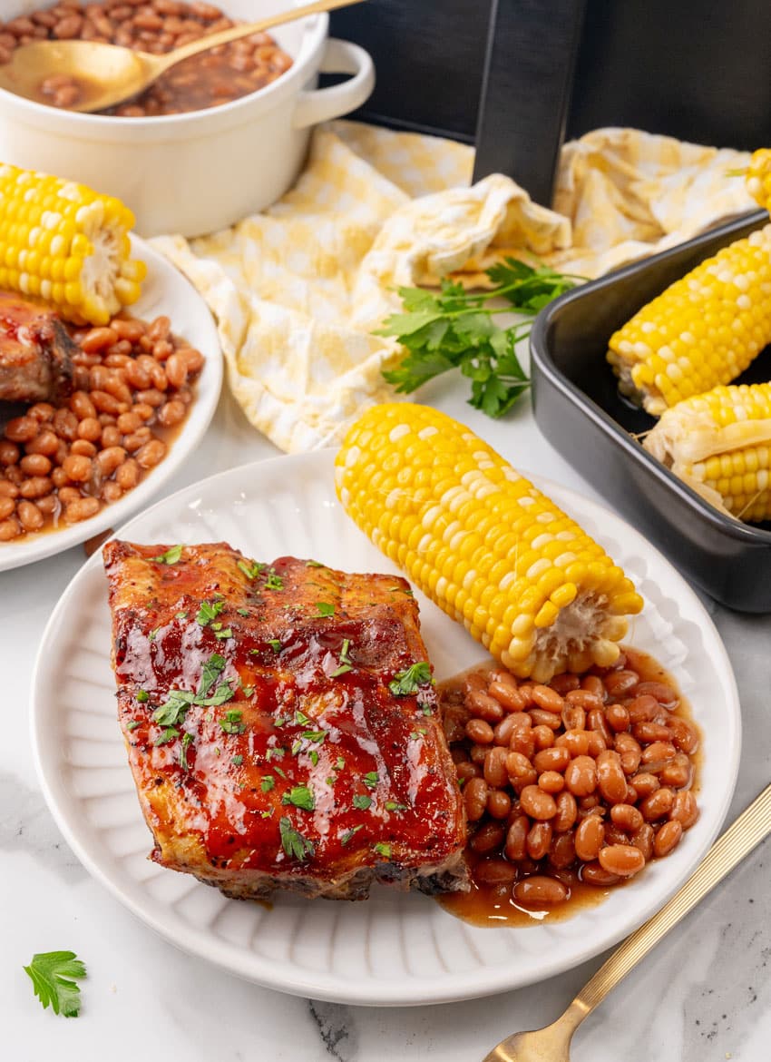 air fryer ribs next to baked beans and corn on the cob on a white plate
