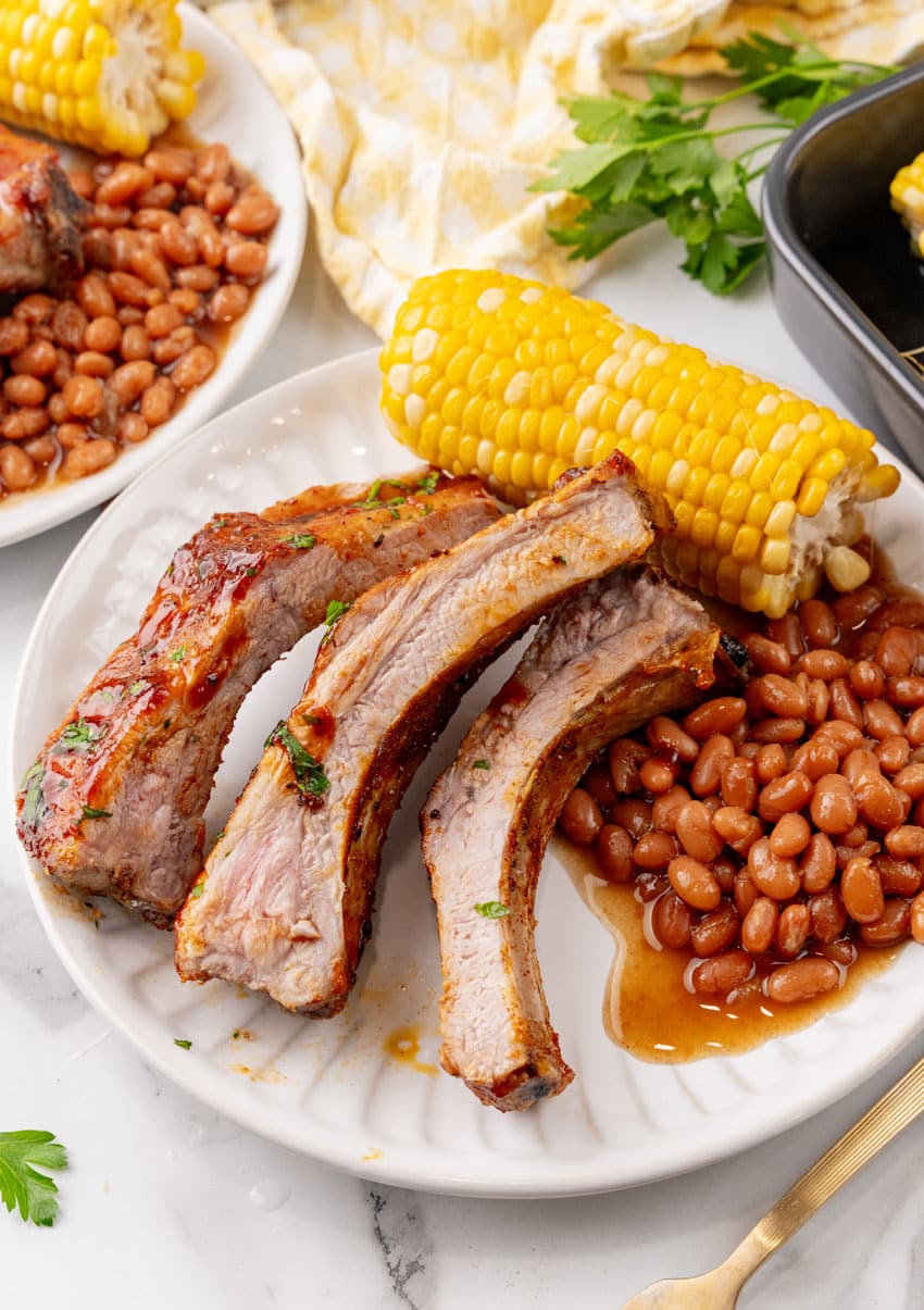 air fryer ribs next to baked beans and corn on the cob on a white plate