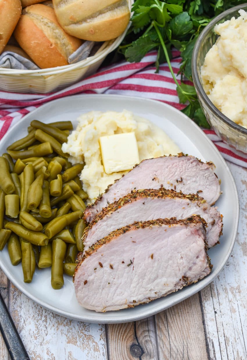 slices of Italian porchetta next to green beans and mashed potatoes on a white plate