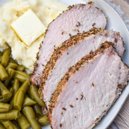 slices of Italian porchetta next to green beans and mashed potatoes on a white plate