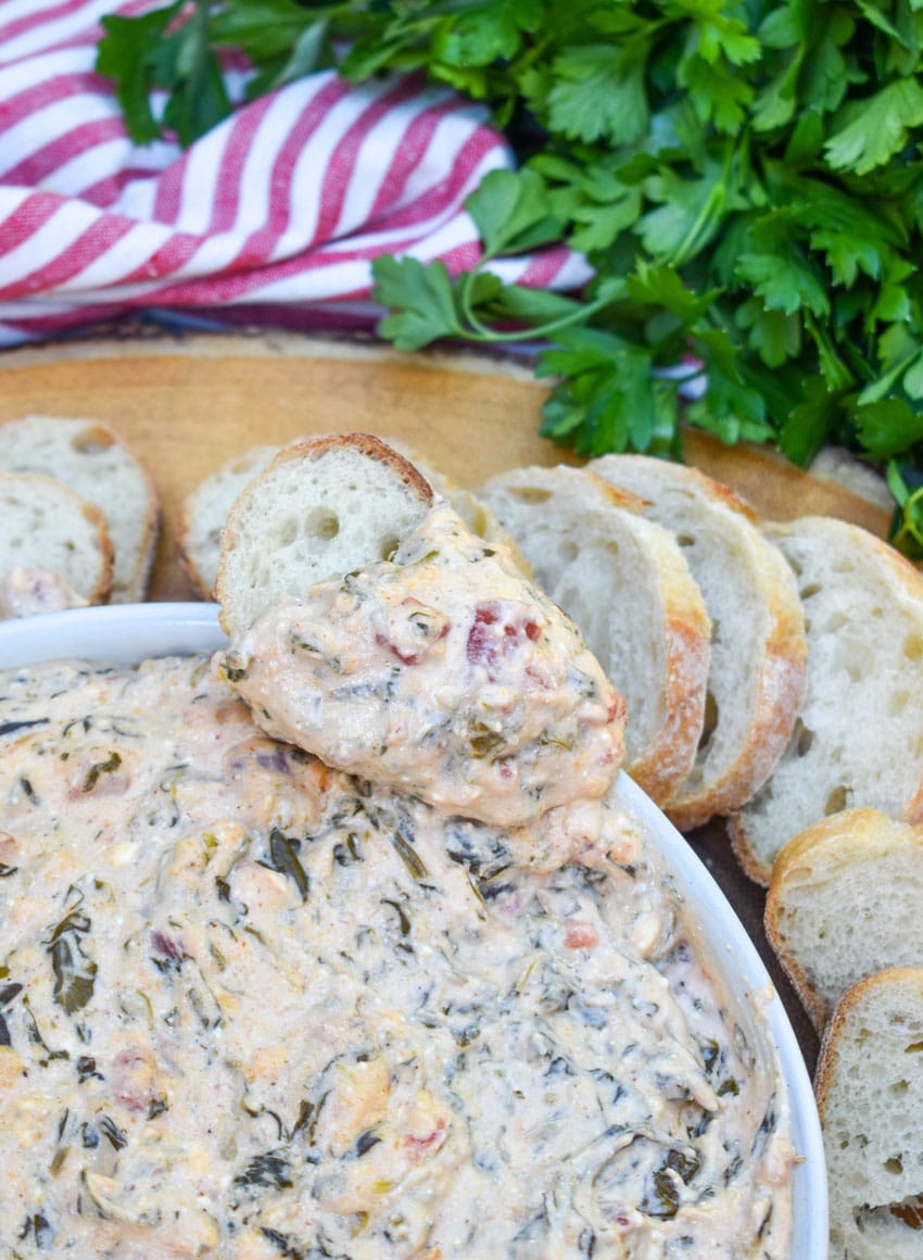 a slice of baguette bread topped with slow cooker spinach dip resting on the edge of a white serving bowl