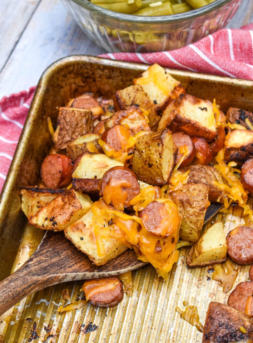 a wooden spatula scooping cheesy hot dogs and potatoes off a metal sheet pan