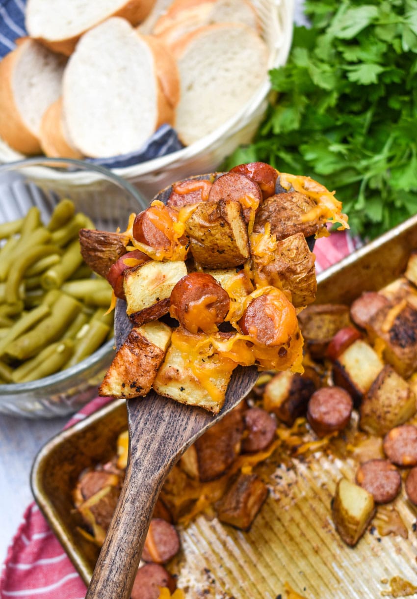 a wooden spatula holding up a scoop of cheesy sheet pan hot dogs and potatoes