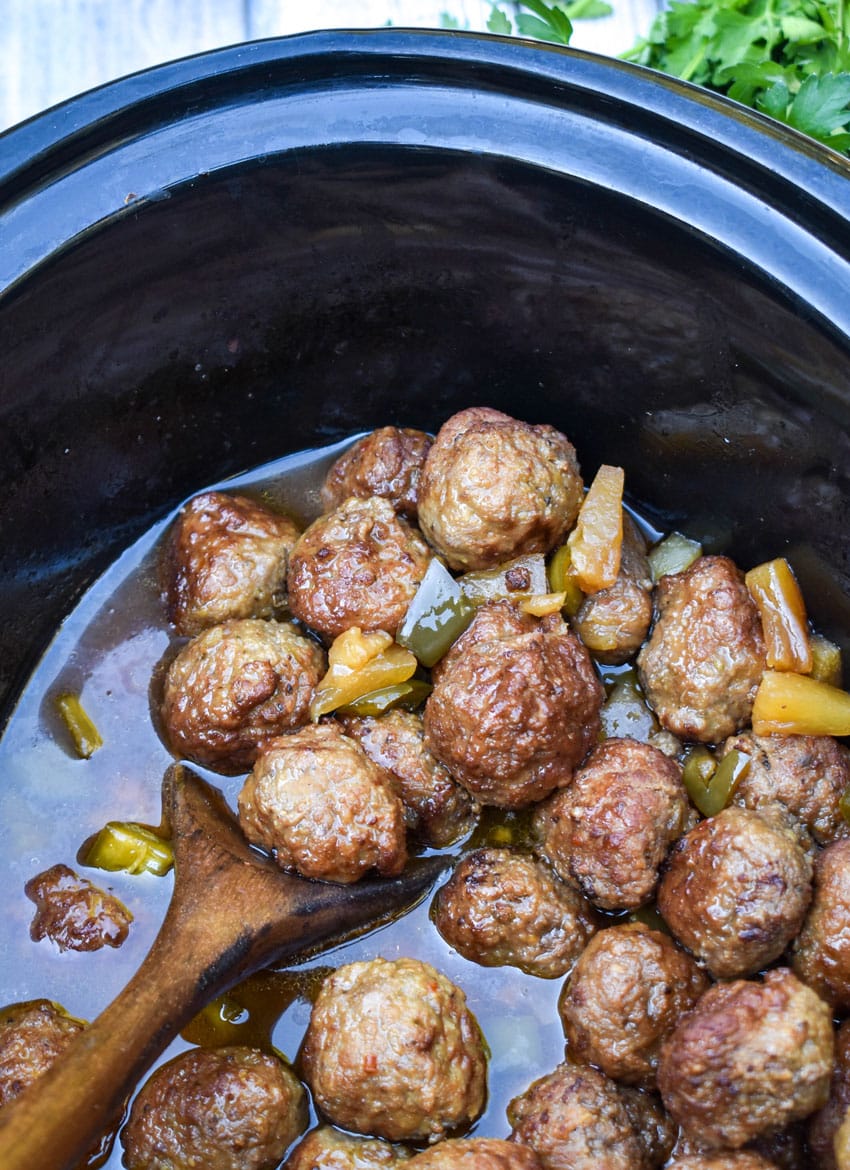 a wooden spoon scooping sweet and sour meatballs out of a black crock pot