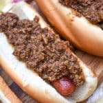 spicy hot dog meat sauce on top of cooked hot dogs in fluffy buns arranged in a row on a wooden cutting board