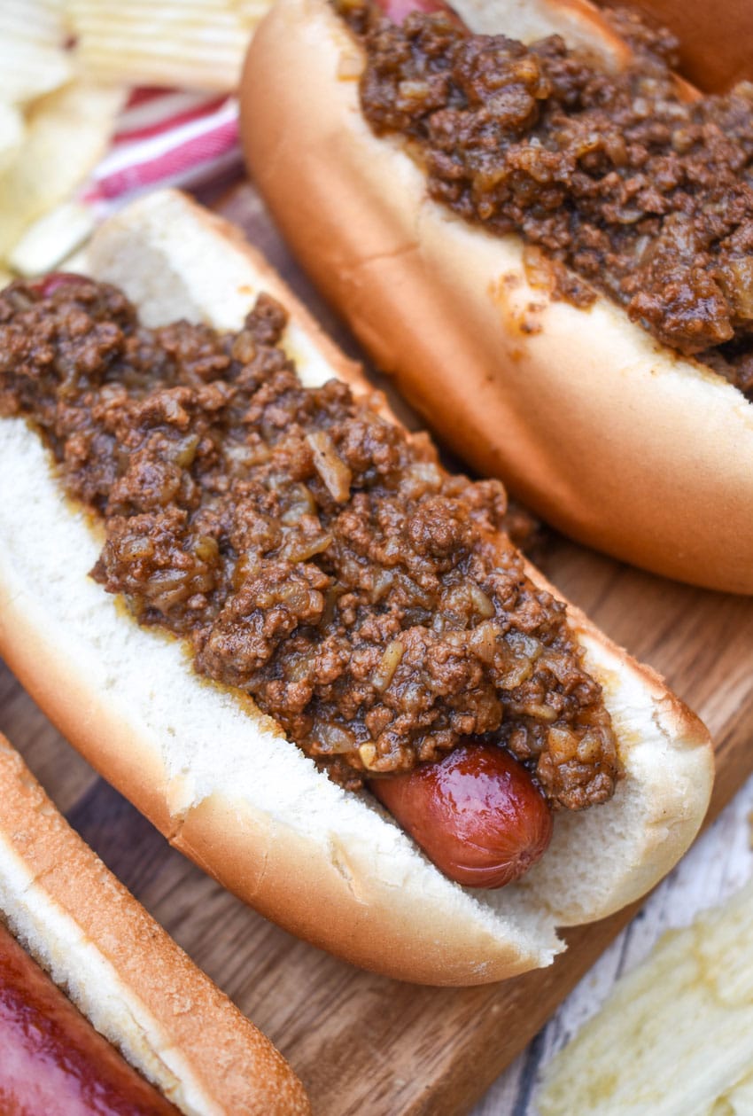 spicy hot dog meat sauce on top of cooked hot dogs in fluffy buns arranged in a row on a wooden cutting board