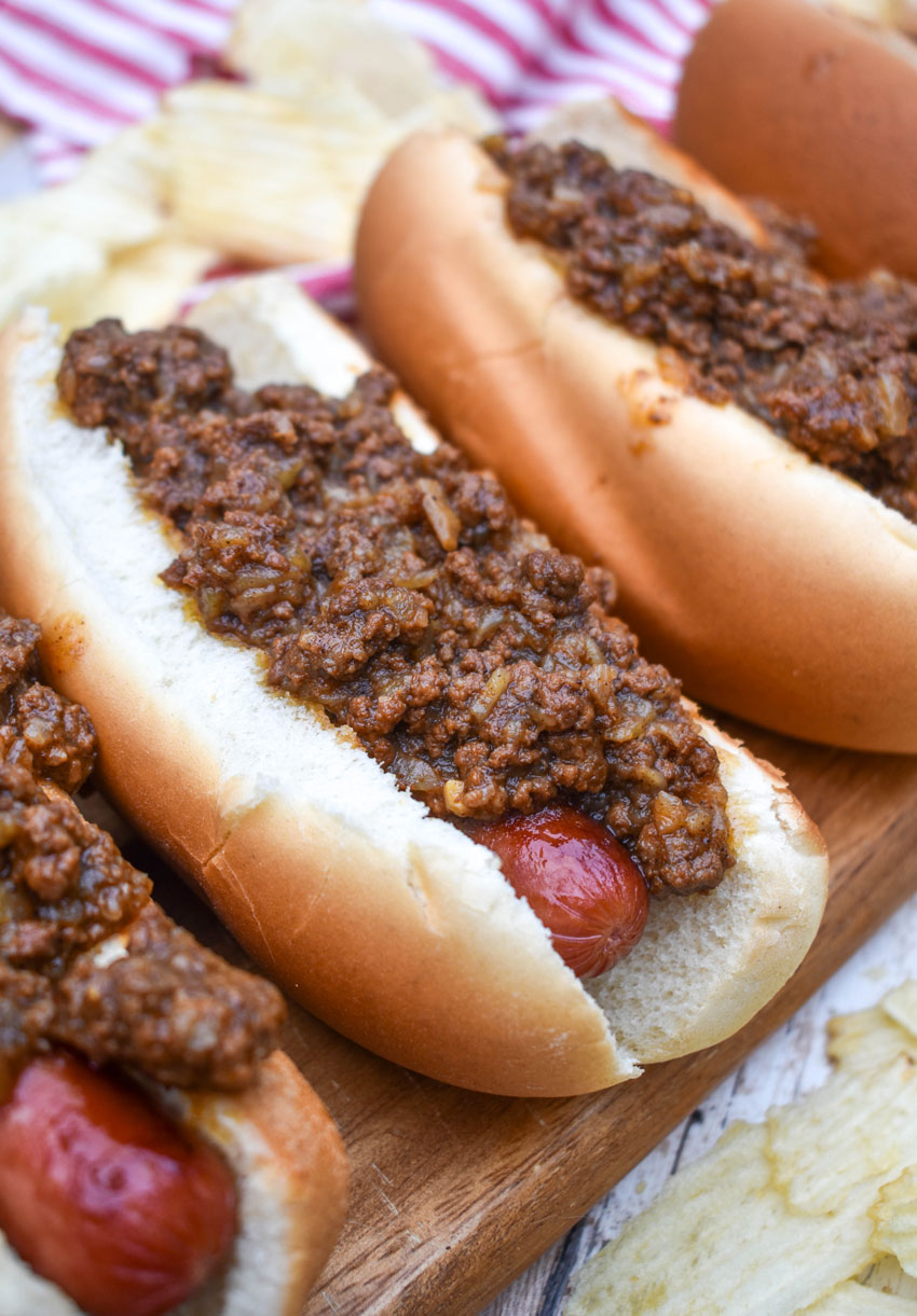 spicy hot dog meat sauce on top of cooked hot dogs in fluffy buns arranged in a row on a wooden cutting board