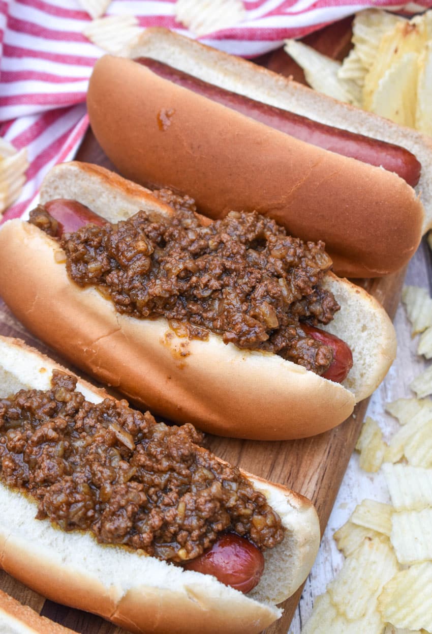 spicy hot dog meat sauce on top of cooked hot dogs in fluffy buns arranged in a row on a wooden cutting board