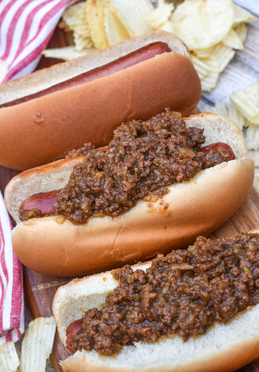 spicy hot dog meat sauce on top of cooked hot dogs in fluffy buns arranged in a row on a wooden cutting board