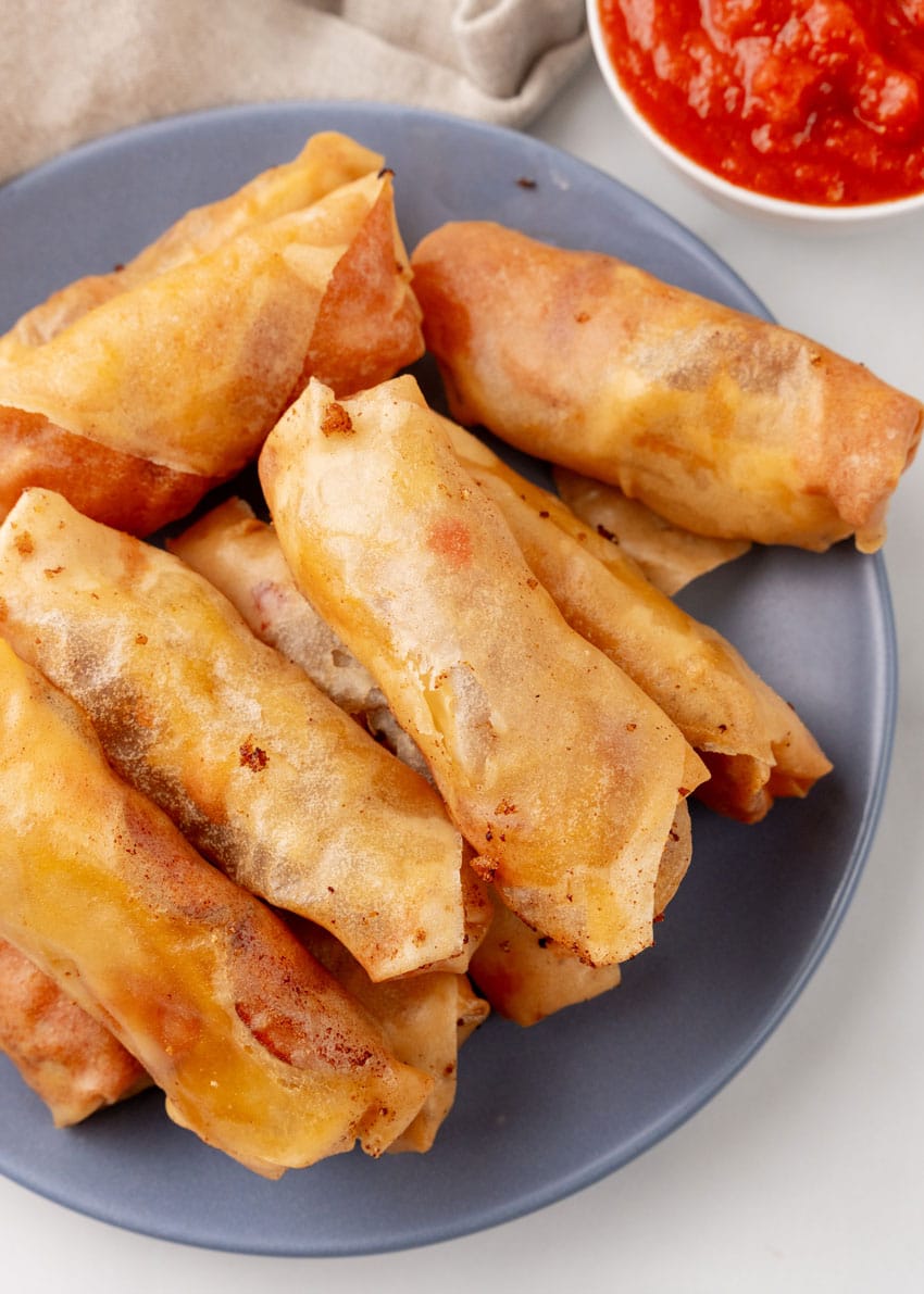 a stack of pepperoni spring rolls on a gray plate with a cup of marinara sauce on the side for dipping