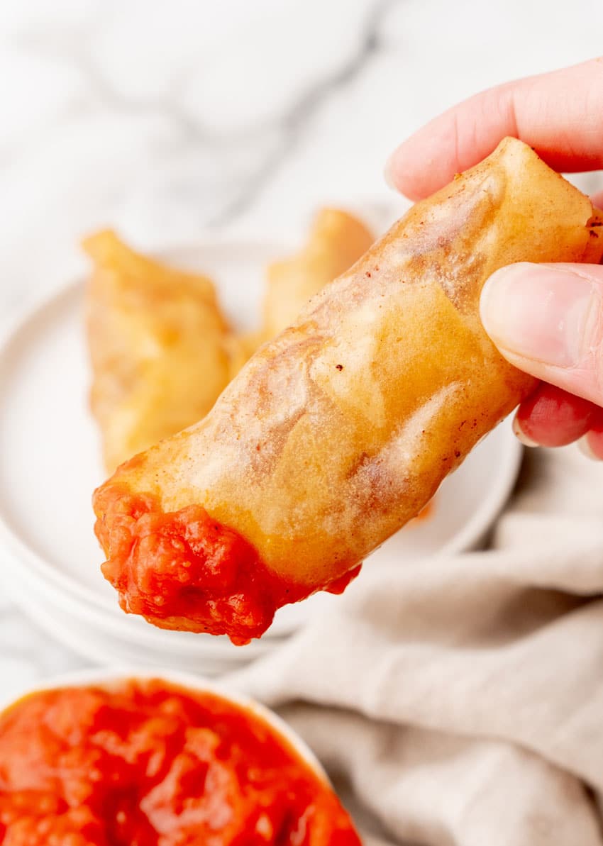 a hand holding up a pepperoni spring roll dipped in marinara sauce