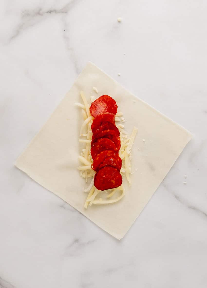 shredded mozzarella cheese and slices of pepperoni arranged in a row on the center of a spring roll wrapper