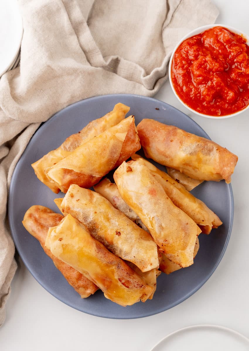 a stack of pepperoni spring rolls on a gray plate with a cup of marinara sauce on the side for dipping