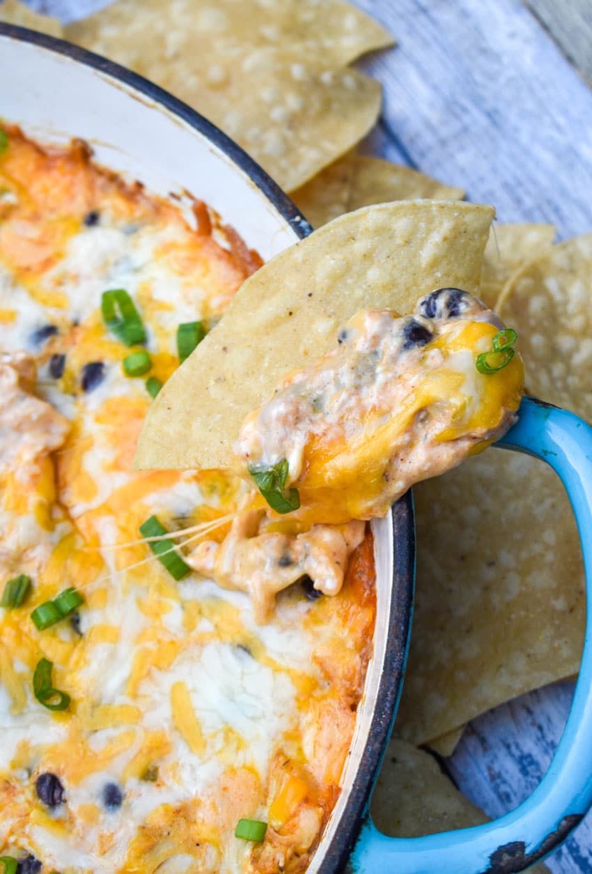 a tortilla chip topped with cheesy chicken enchilada dip resting on the edge of a cast iron skillet