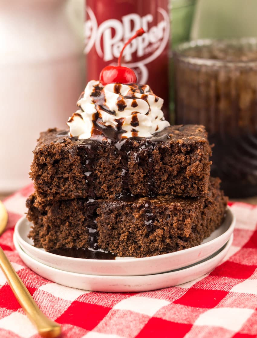 two dr pepper brownies topped with whipped cream and chocolate sauce stacked on a small white plate