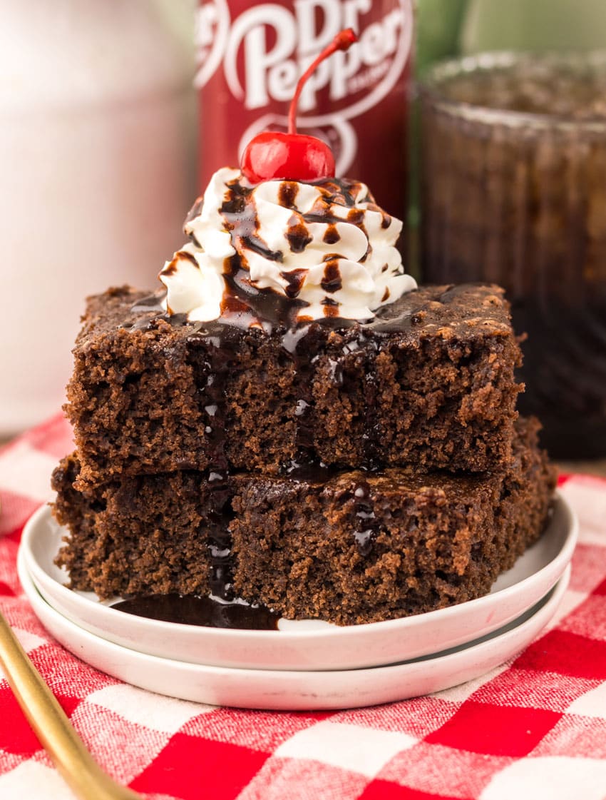 two dr pepper brownies topped with whipped cream and chocolate sauce stacked on a small white plate