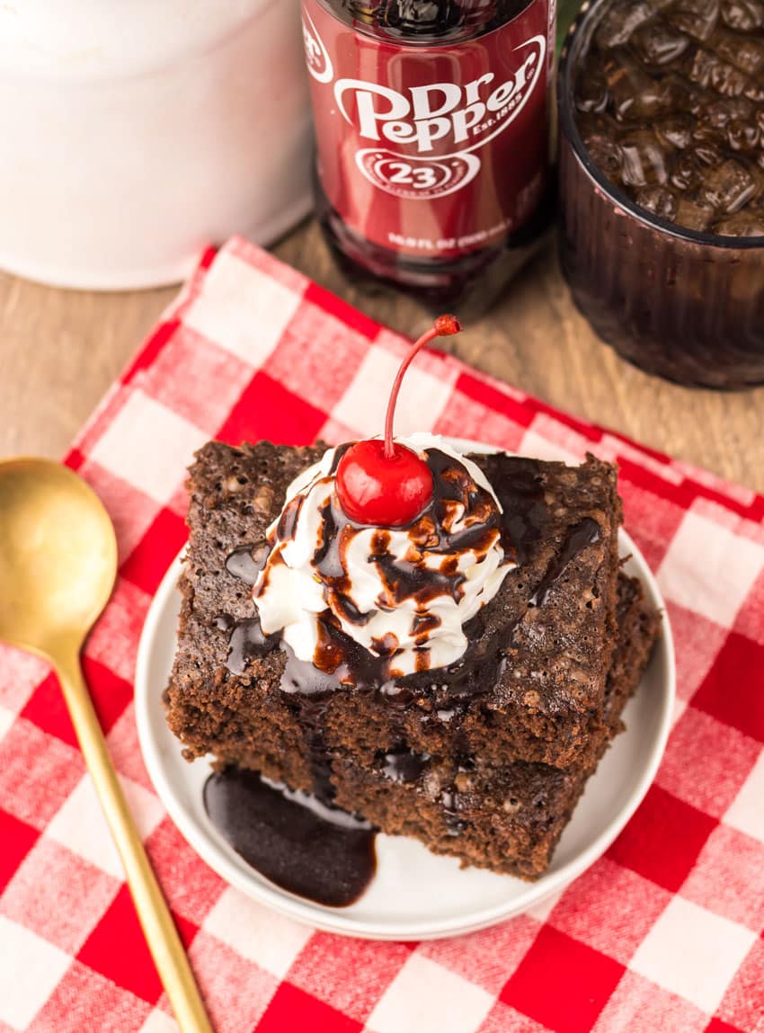 two dr pepper brownies topped with whipped cream and chocolate sauce stacked on a small white plate