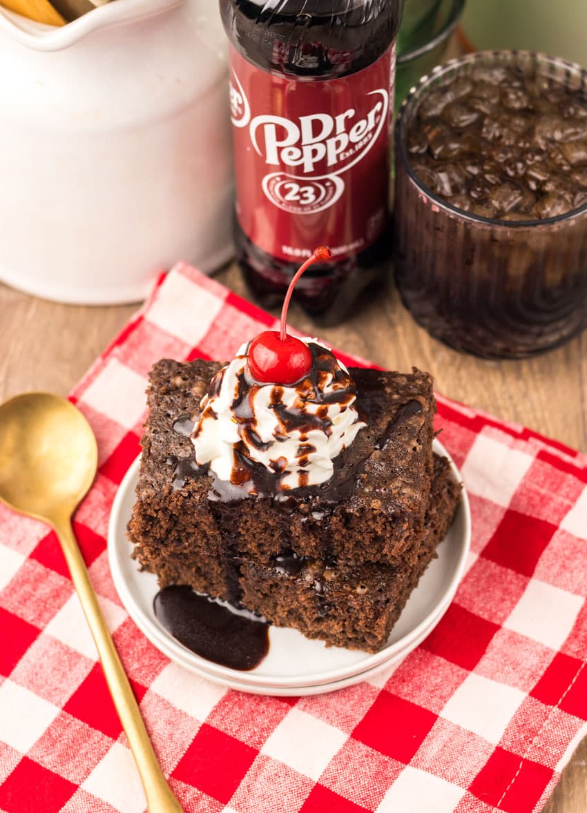 two dr pepper brownies topped with whipped cream and chocolate sauce stacked on a small white plate