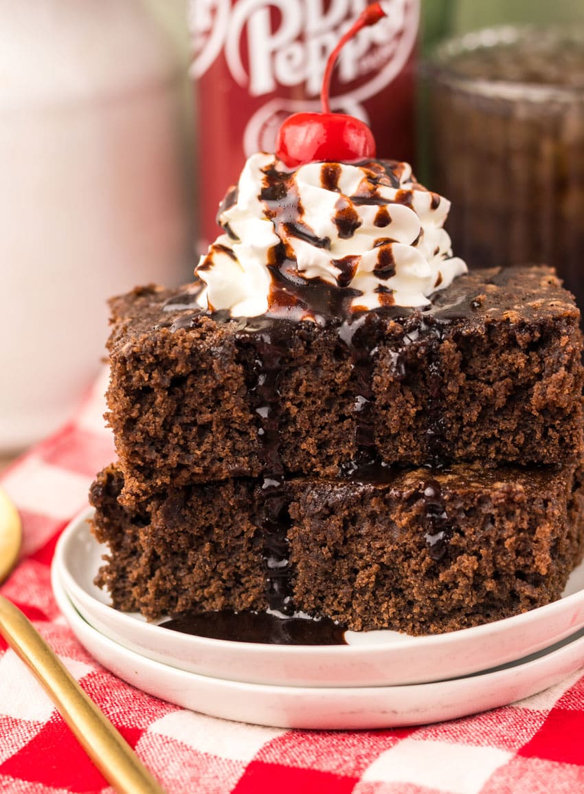 two dr pepper brownies topped with whipped cream and chocolate sauce stacked on a small white plate