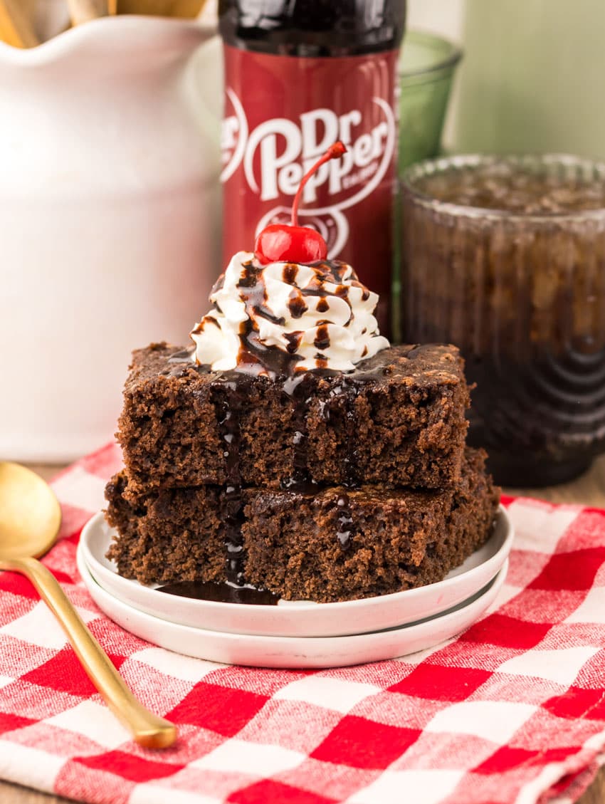 two dr pepper brownies topped with whipped cream and chocolate sauce stacked on a small white plate