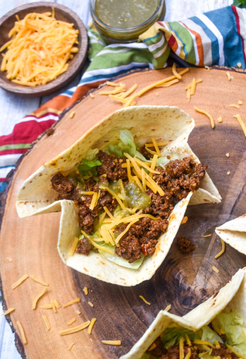 homemade flour tortilla bowls filled with chopped lettuce, seasoned ground beef, and shredded cheese on a wooden cutting board