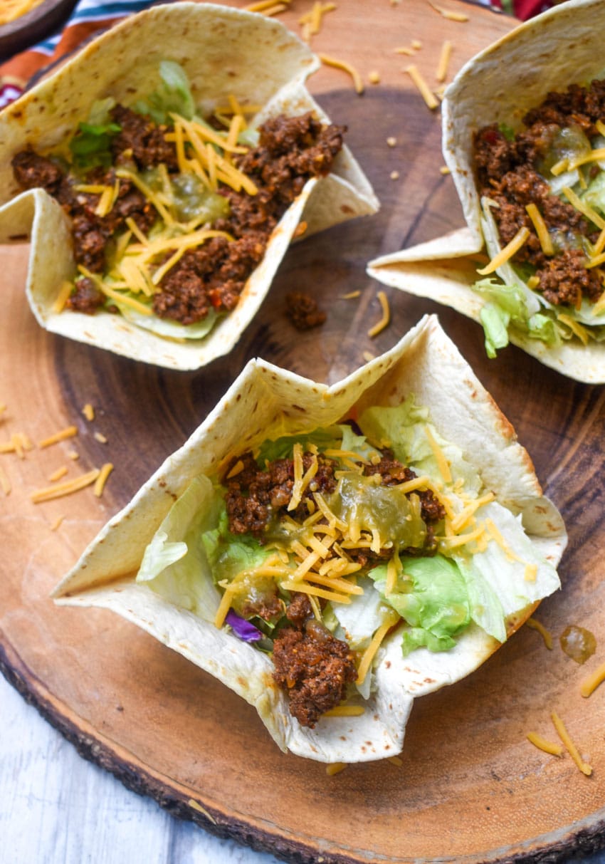 homemade flour tortilla bowls filled with chopped lettuce, seasoned ground beef, and shredded cheese on a wooden cutting board