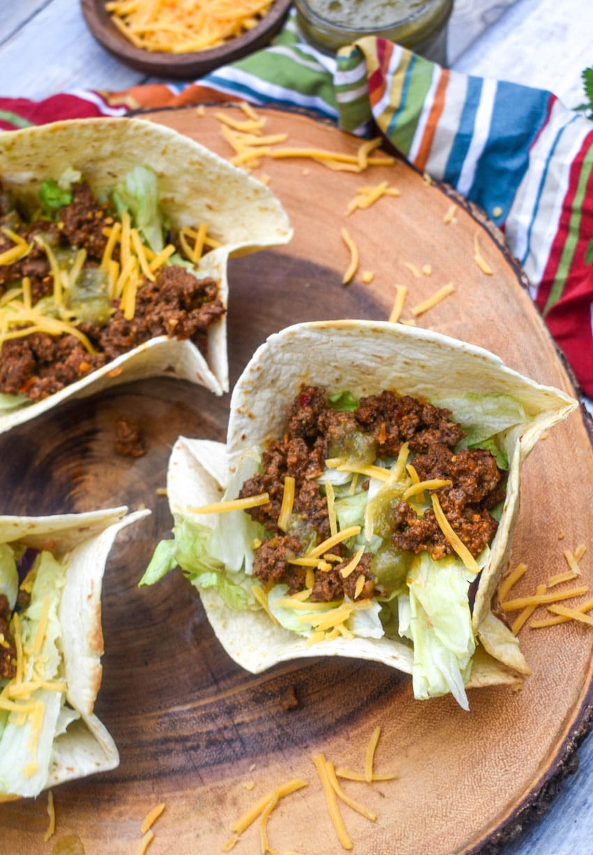 homemade flour tortilla bowls filled with chopped lettuce, seasoned ground beef, and shredded cheese on a wooden cutting board