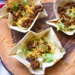 homemade flour tortilla bowls filled with chopped lettuce, seasoned ground beef, and shredded cheese on a wooden cutting board