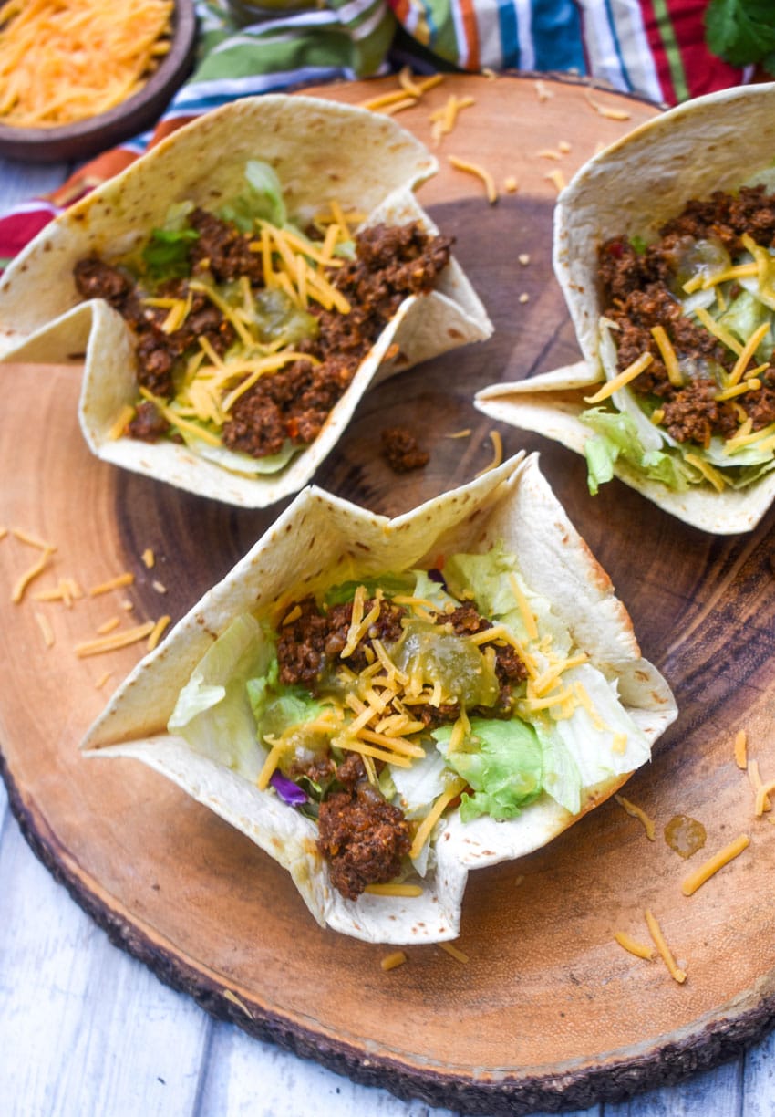 homemade flour tortilla bowls filled with chopped lettuce, seasoned ground beef, and shredded cheese on a wooden cutting board