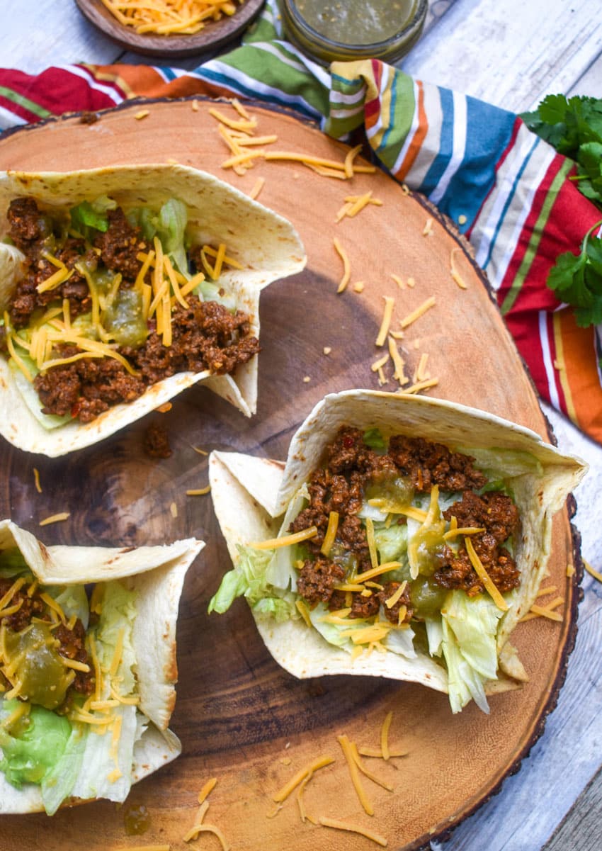 homemade flour tortilla bowls filled with chopped lettuce, seasoned ground beef, and shredded cheese on a wooden cutting board