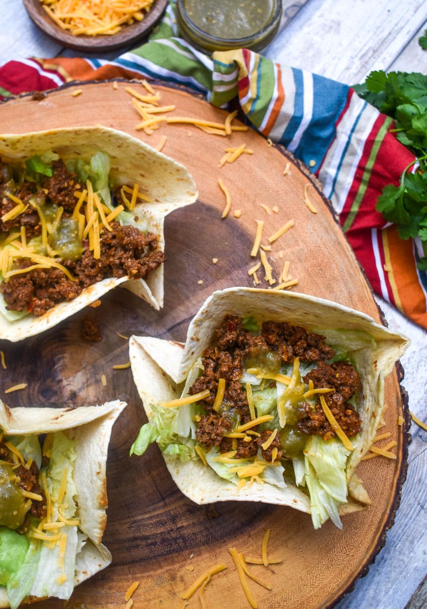 homemade flour tortilla bowls filled with chopped lettuce, seasoned ground beef, and shredded cheese on a wooden cutting board