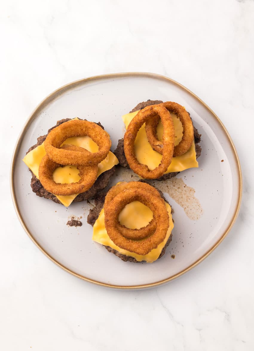 two grilled hamburger patties topped with slices of American cheese and crispy onion rings on a gray plate