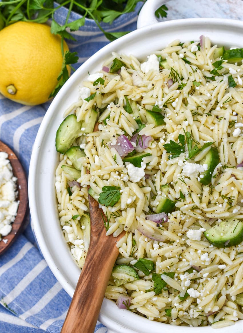 a wooden spoon scooping cucumber lemon orzo salad out of a white serving bowl