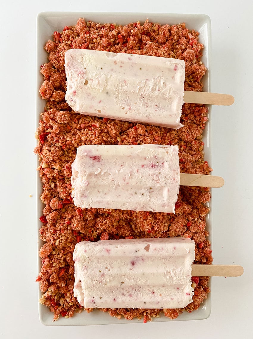 homemade strawberry ice cream popsicles laying on strawberry crunch topping on a white platter