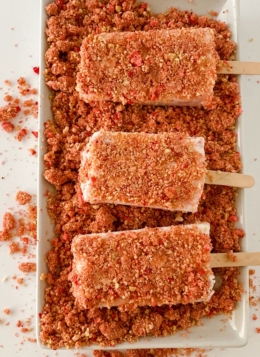 homemade strawberry ice cream popsicles laying on strawberry crunch topping on a white platter