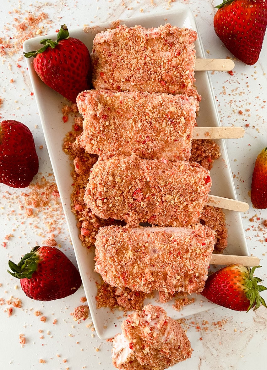 homemade strawberry ice cream popsicles laying on strawberry crunch topping on a white platter