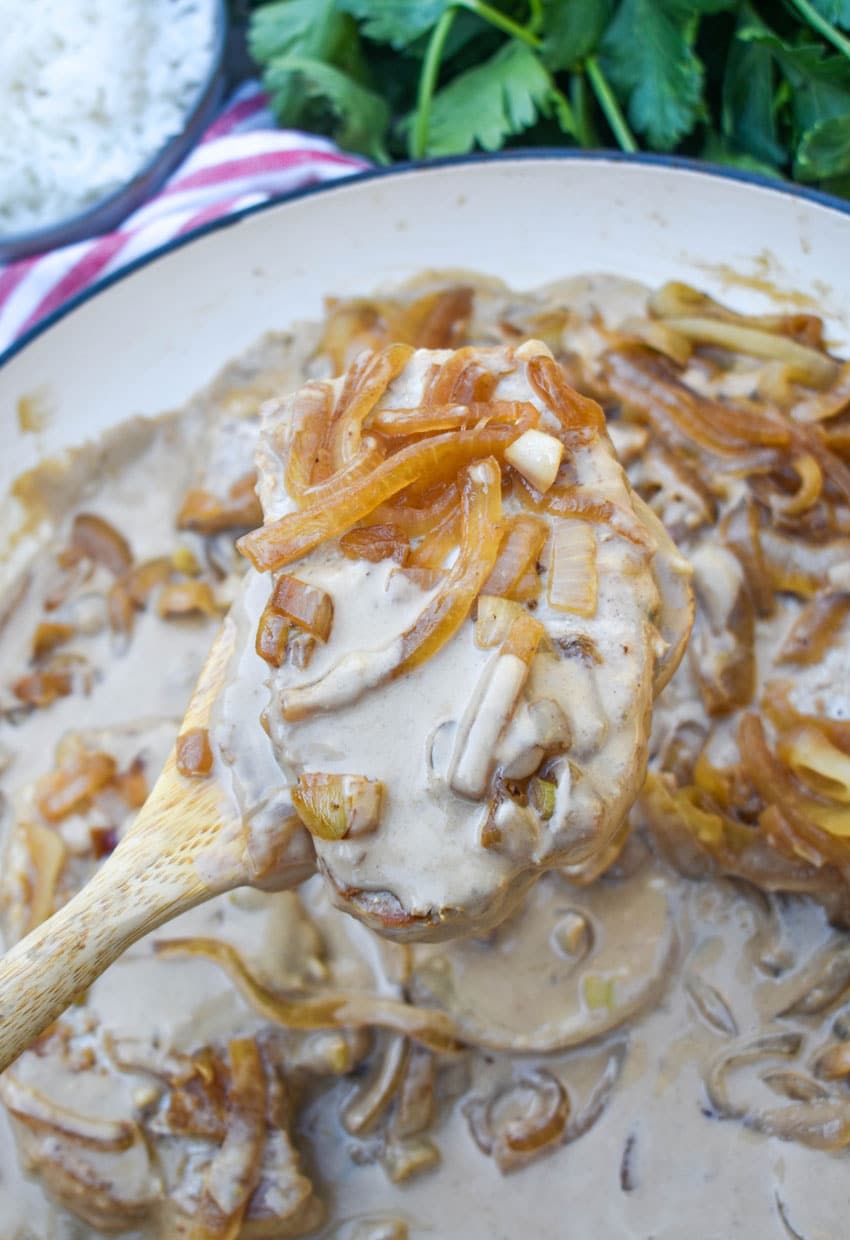 a wooden spatula holding up a pork chop in cream sauce topped with caramelized onions