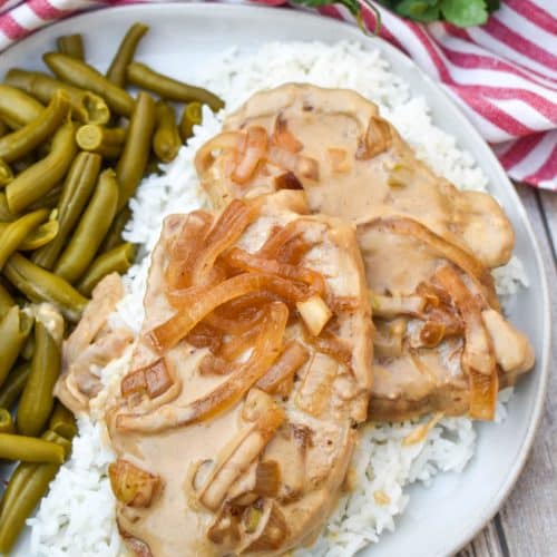 pork chops with cream sauce and onions over steamed white rice next to green beans on a white dinner plate